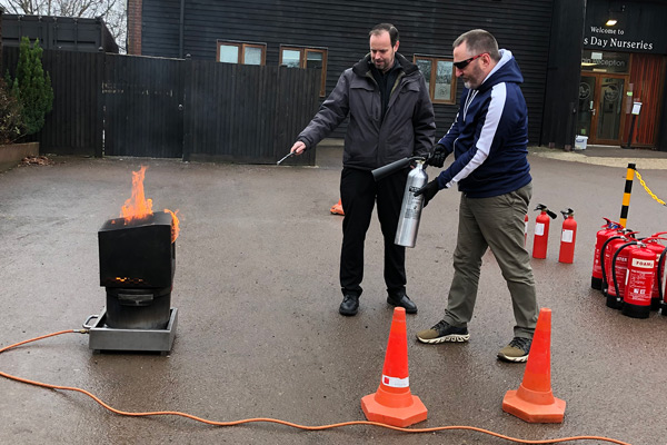 staff fire training image, two staff members facing a flaming object outdoors with fire extinguishers